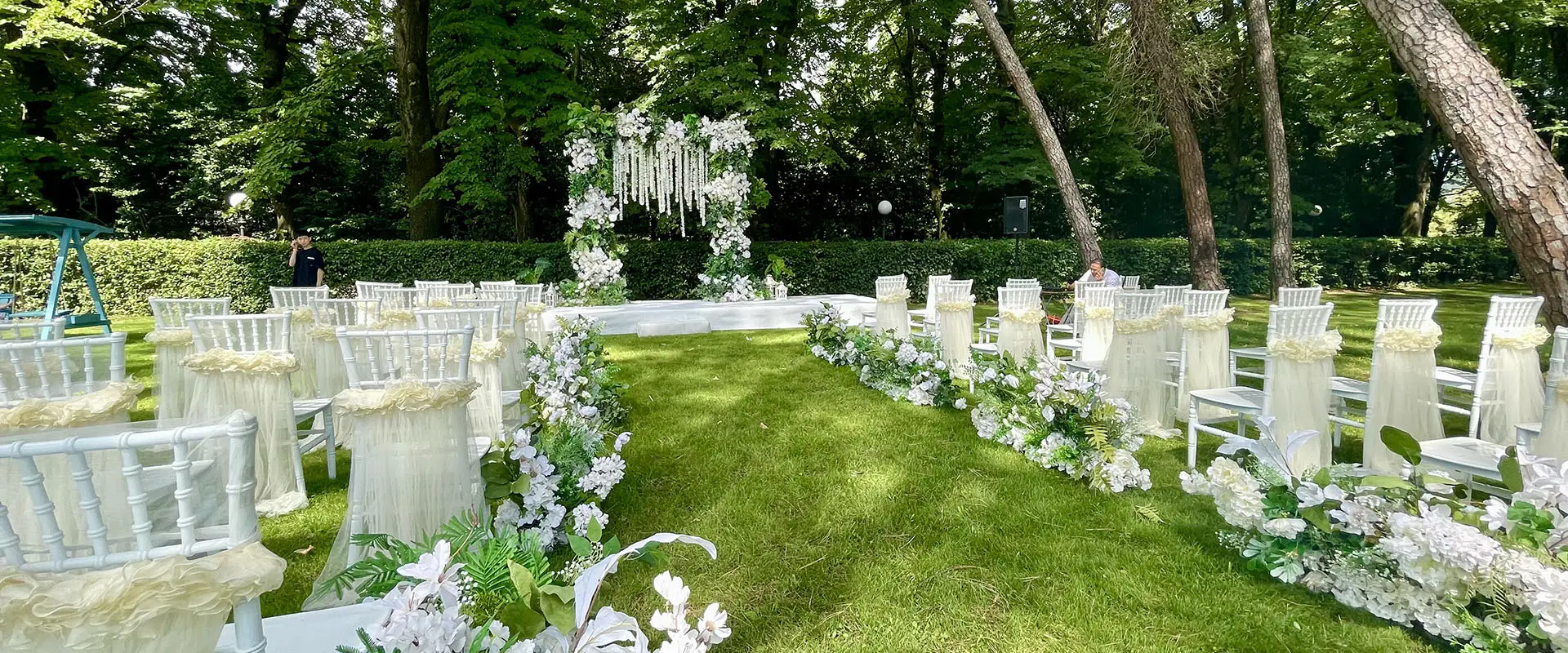 Il parco allestito per una cerimonia: le sedie bianche ai lati di un corridoio di fiori, con la composizione sospesa in fondo
