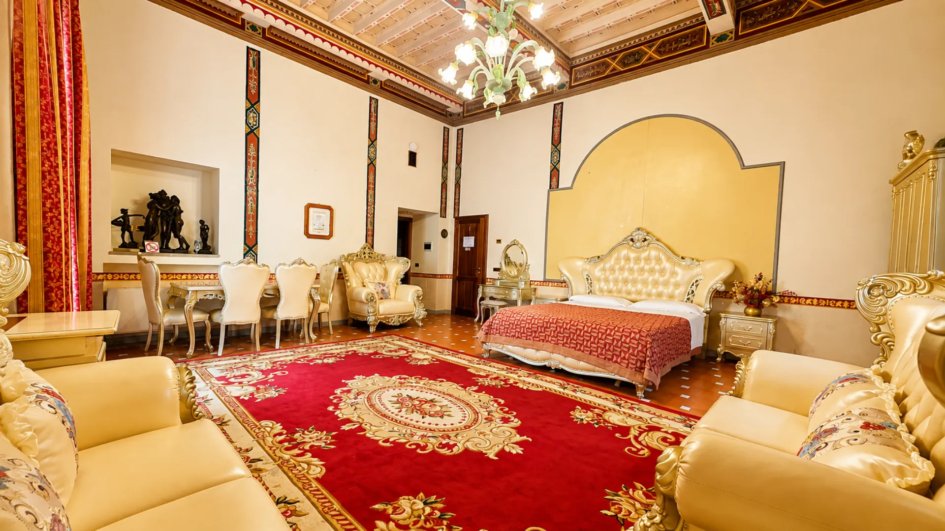 La Junior Suite vista dall'ingresso: soffitto a travi dipinte, lampadario di vetro, tavolo da pranzo e letto matrimoniale in fondo
