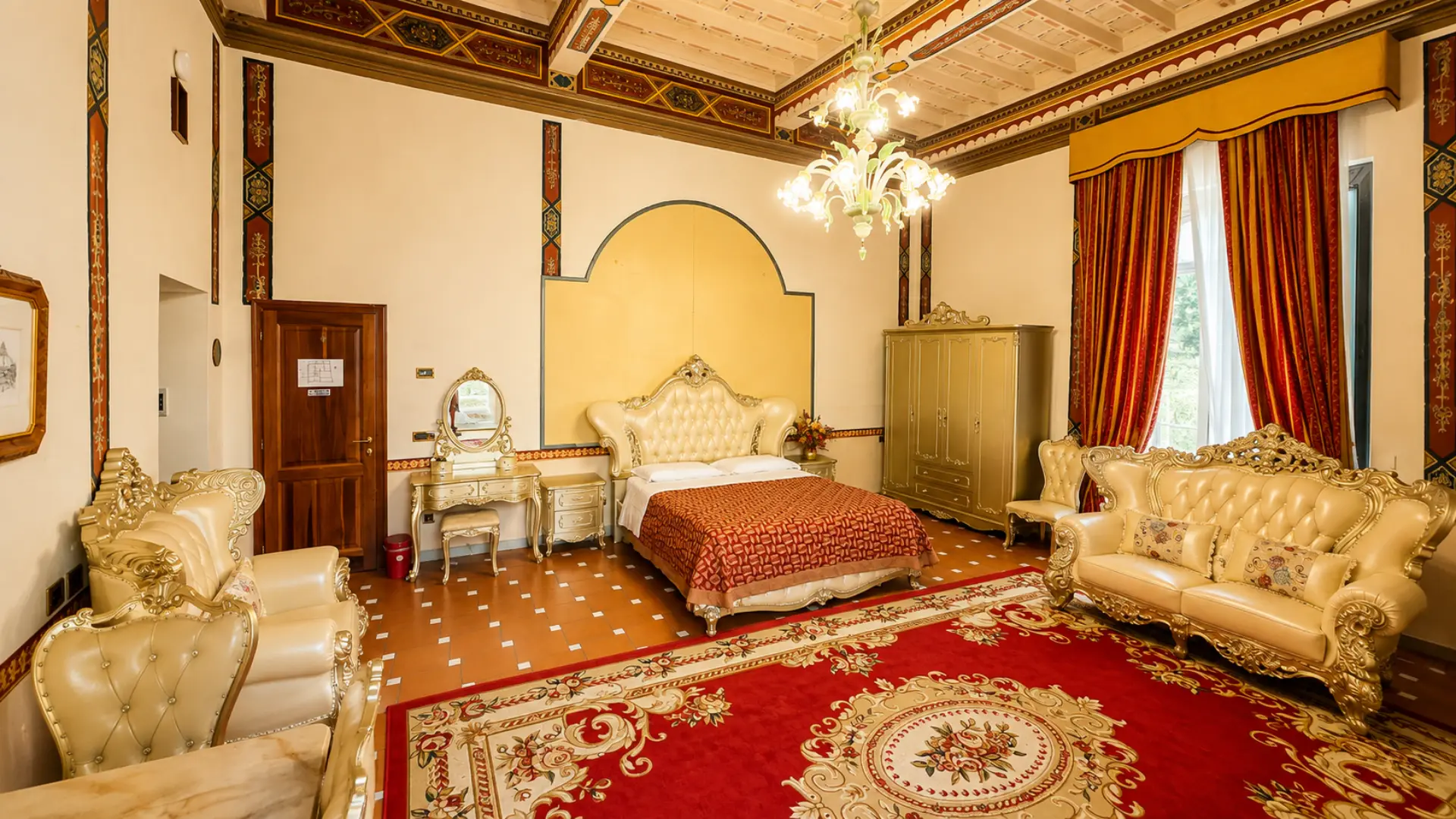 Il letto capitonné della Junior Suite fra la toeletta e l'armadio dorato, con il tappeto rosso e le tende damascate