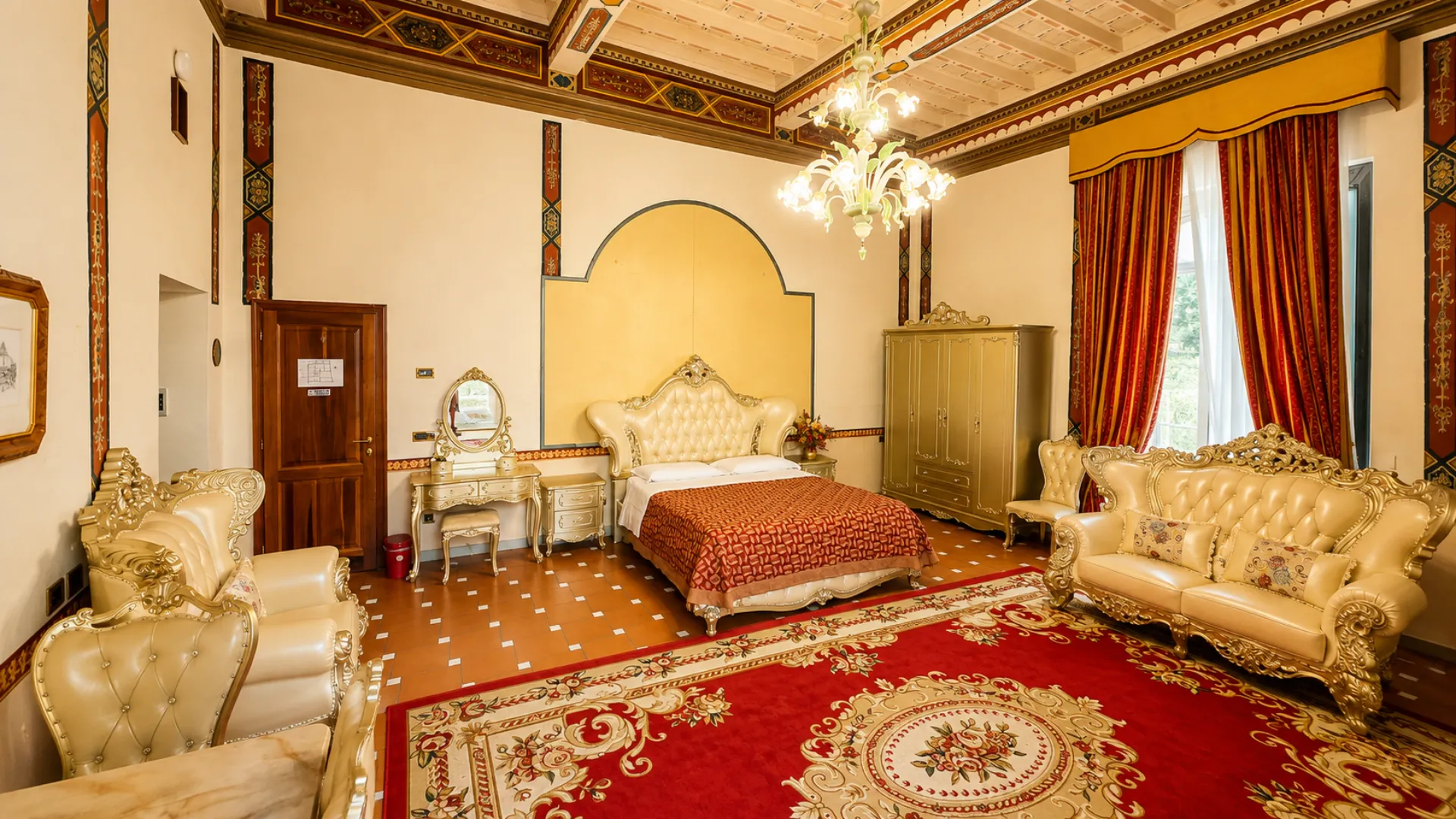 Il letto capitonné della Junior Suite fra la toeletta e l'armadio dorato, con il tappeto rosso e le tende damascate