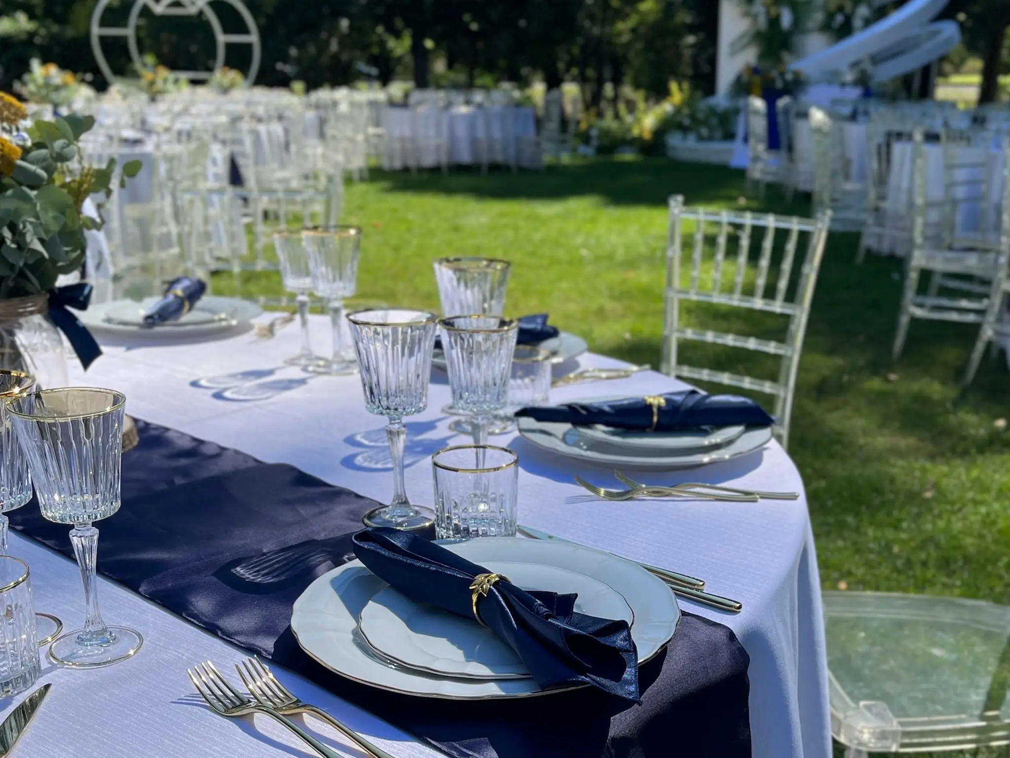 Un matrimonio allestito nel parco della villa: la tavolata apparecchiata sul prato, le sedie trasparenti e l'arco di fiori sullo sfondo