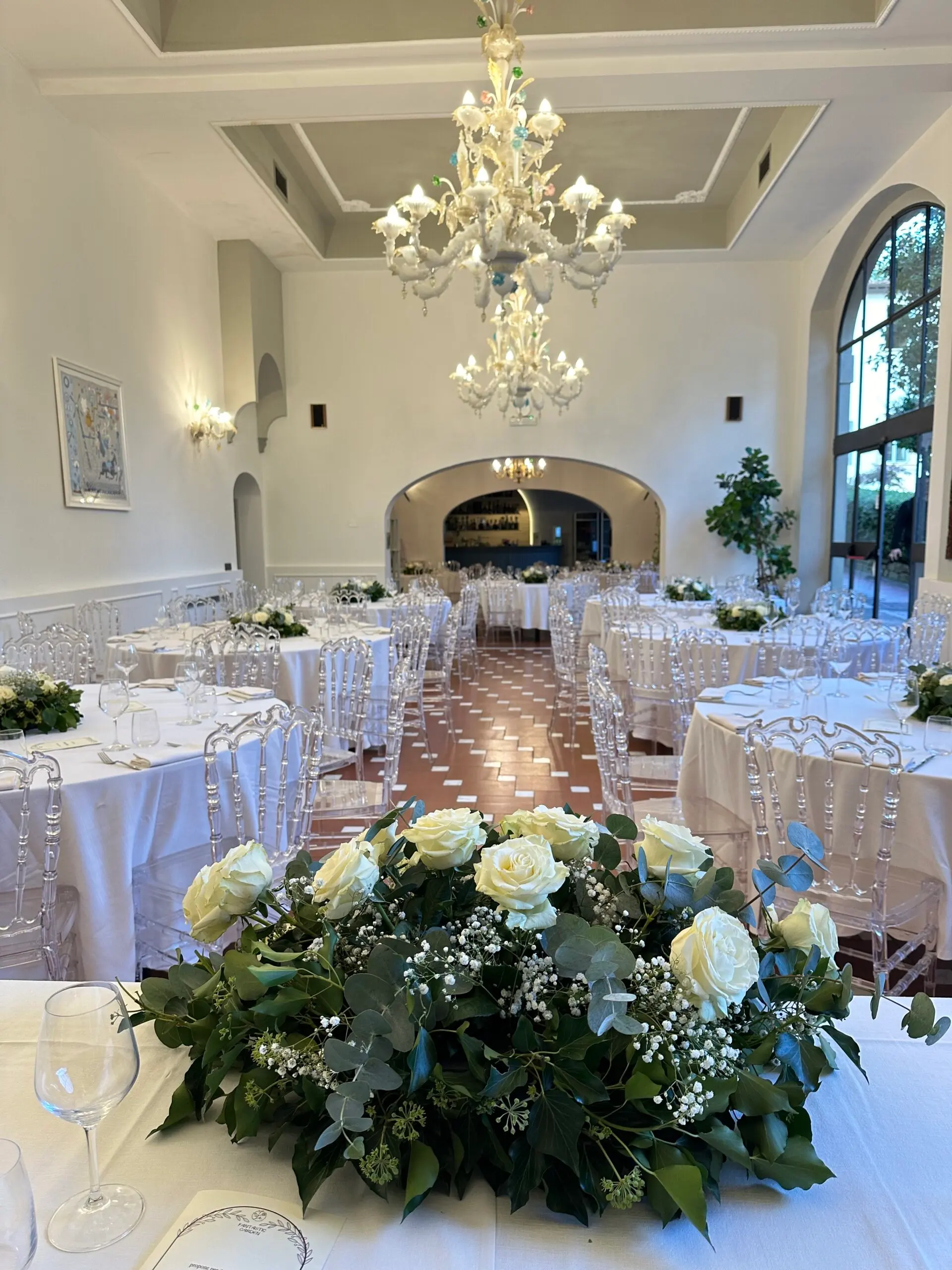 La sala della villa allestita per un matrimonio, con i tavoli bianchi e i centrotavola di rose sotto il lampadario di vetro
