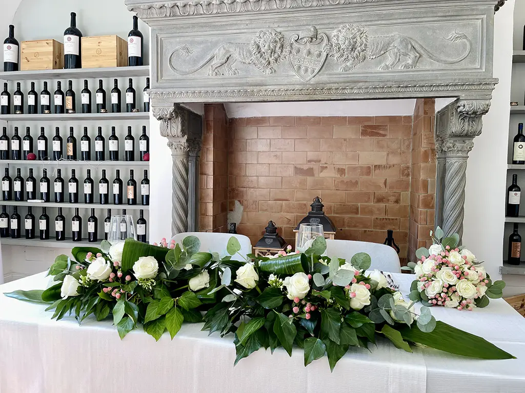 Il tavolo degli sposi davanti al camino in pietra con lo stemma, con la ghirlanda di rose bianche e gli scaffali del vino dietro