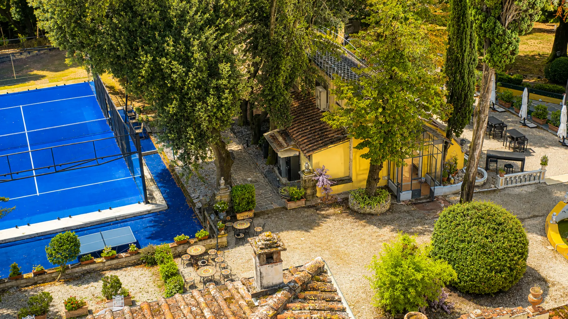 Il campo da padel visto dall'alto: il fondo blu fra gli alberi del parco, i tavolini all'ombra e la limonaia gialla della villa accanto
