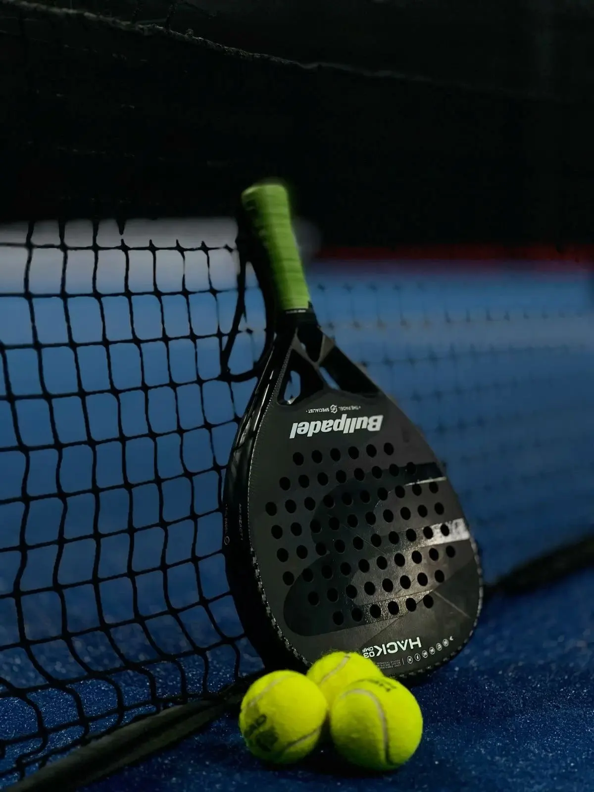 Una racchetta da padel appoggiata alla rete con due palline a terra