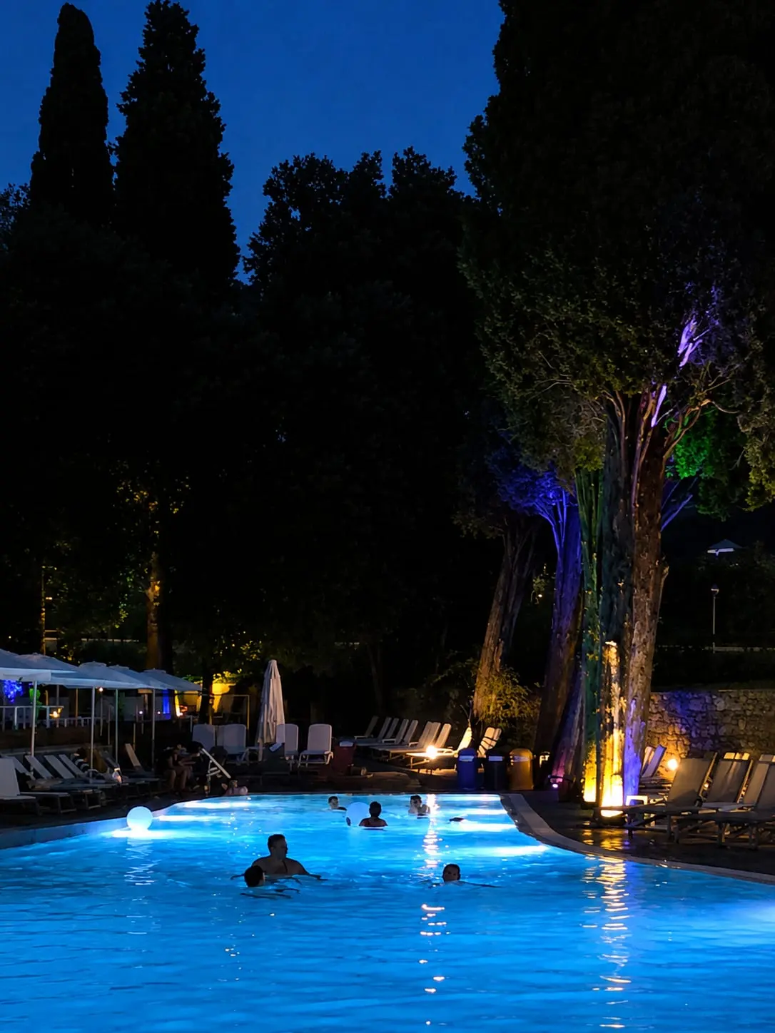 Il bagno serale: gente in acqua nella piscina illuminata di blu, con gli alberi del parco accesi di viola e arancio