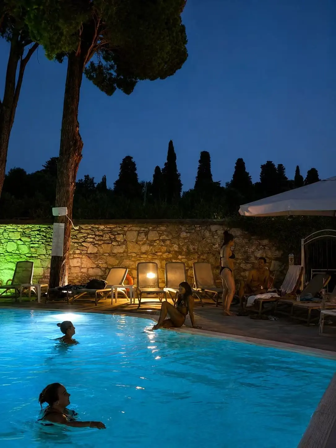 La piscina illuminata di sera, con il muro in pietra e i lettini a bordo vasca