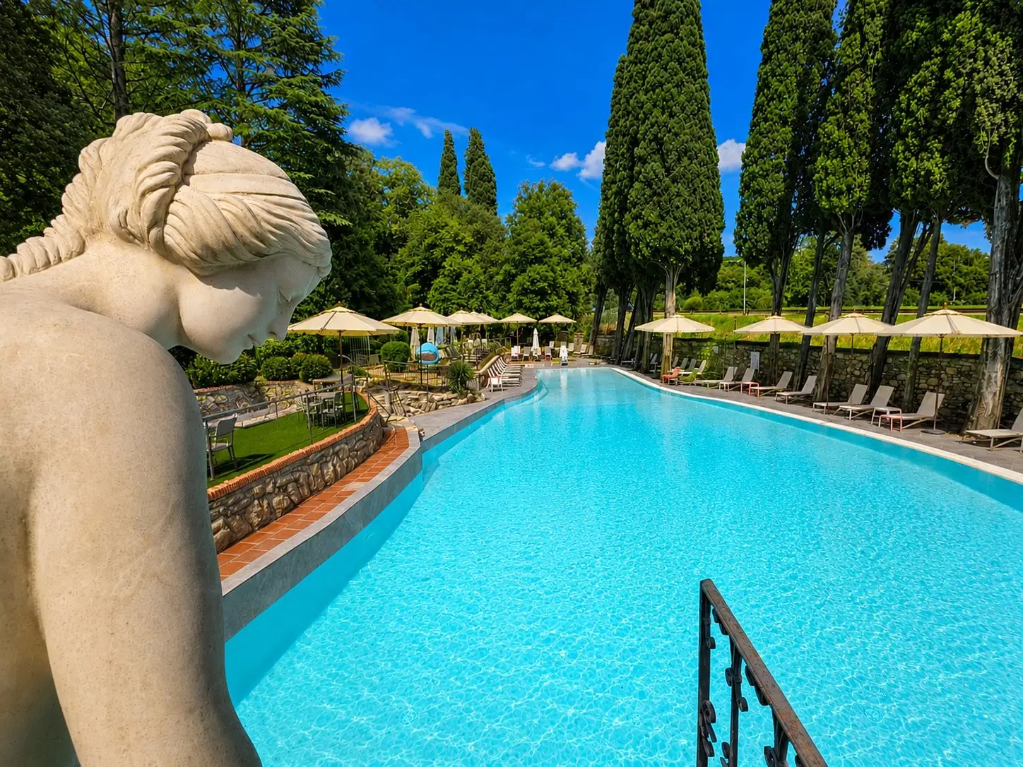 La piscina vista da dietro una statua in pietra, con i cipressi, gli ombrelloni e i lettini lungo il bordo
