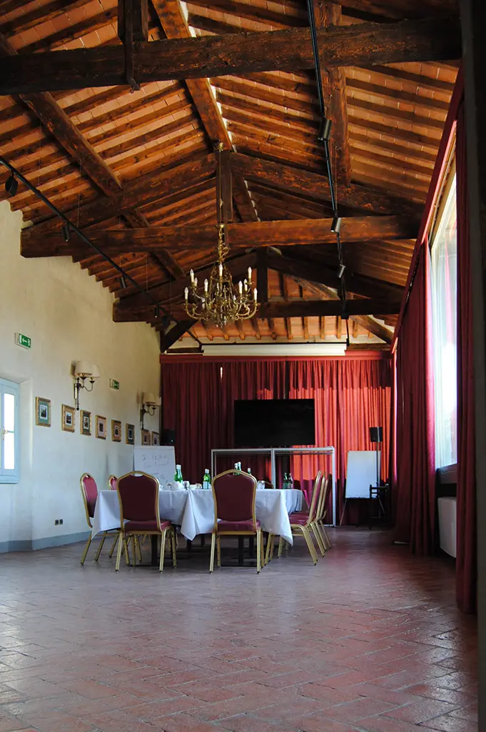 La sala allestita per un corso: tavoli a ferro di cavallo, televisore e lavagna a fogli mobili sotto la volta di travi