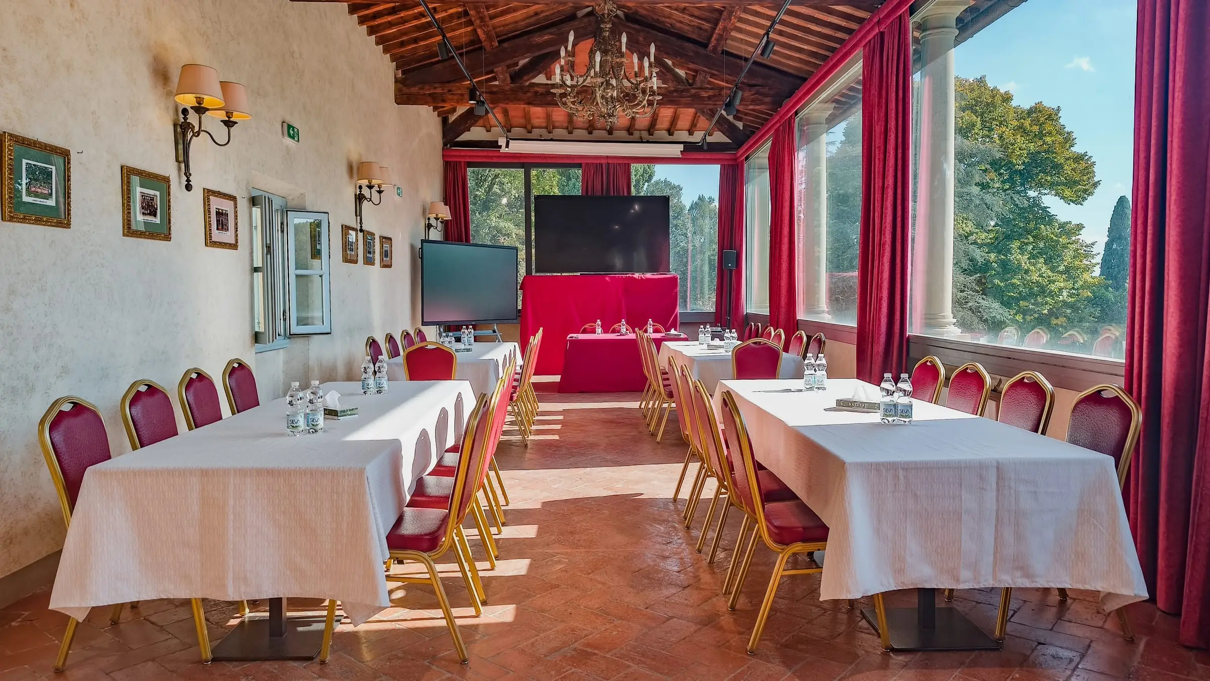 La sala meeting allestita a ferro di cavallo sotto la volta di travi: tavoli con le tovaglie chiare, sedie rosse e oro, il grande schermo in fondo e le vetrate con le tende rosse sul parco
