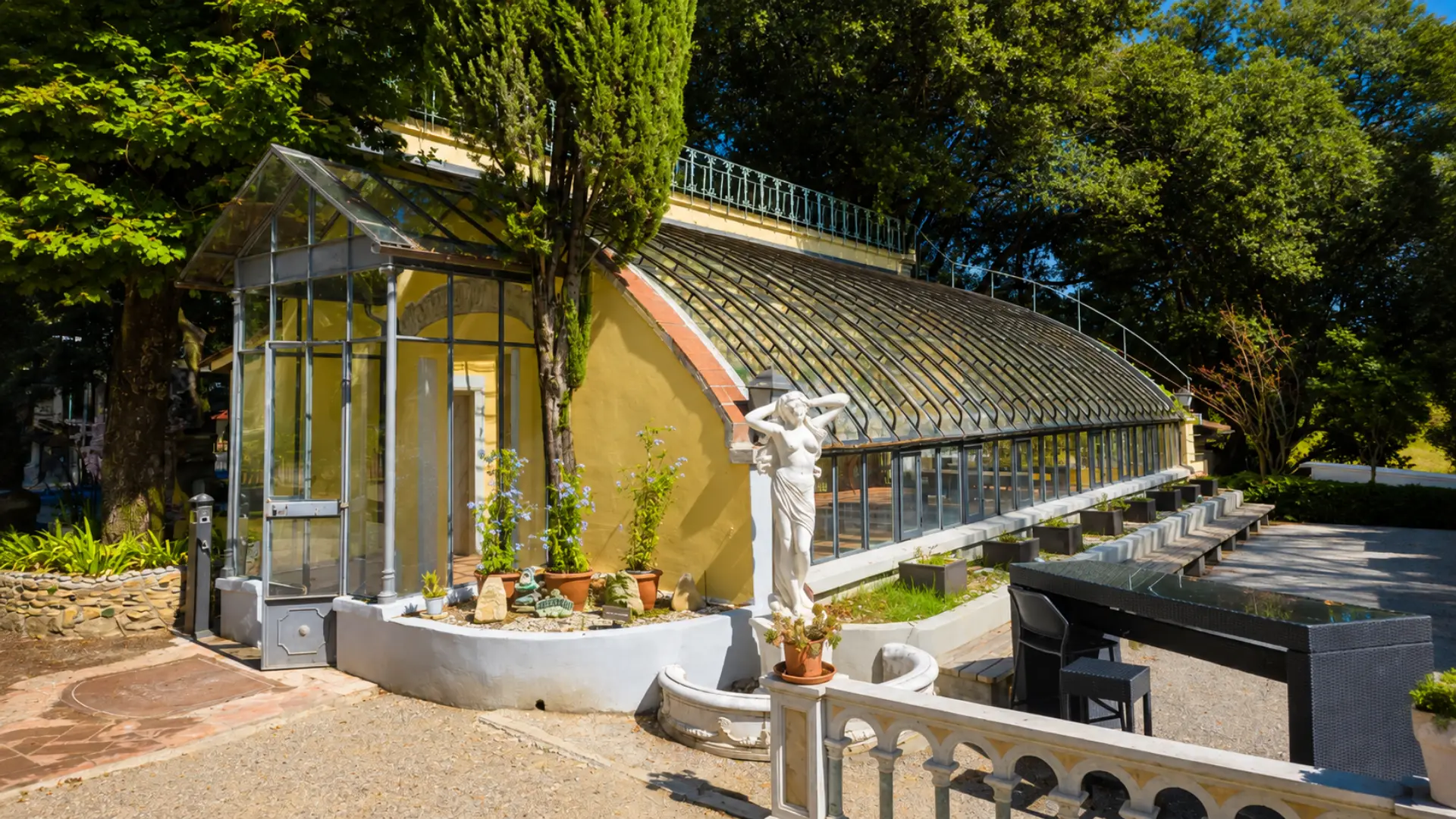 La serra vista dal parco: la lunga volta a vetri sull'edificio giallo ocra, il portico in ferro battuto dell'ingresso e una statua bianca fra i vasi di terracotta