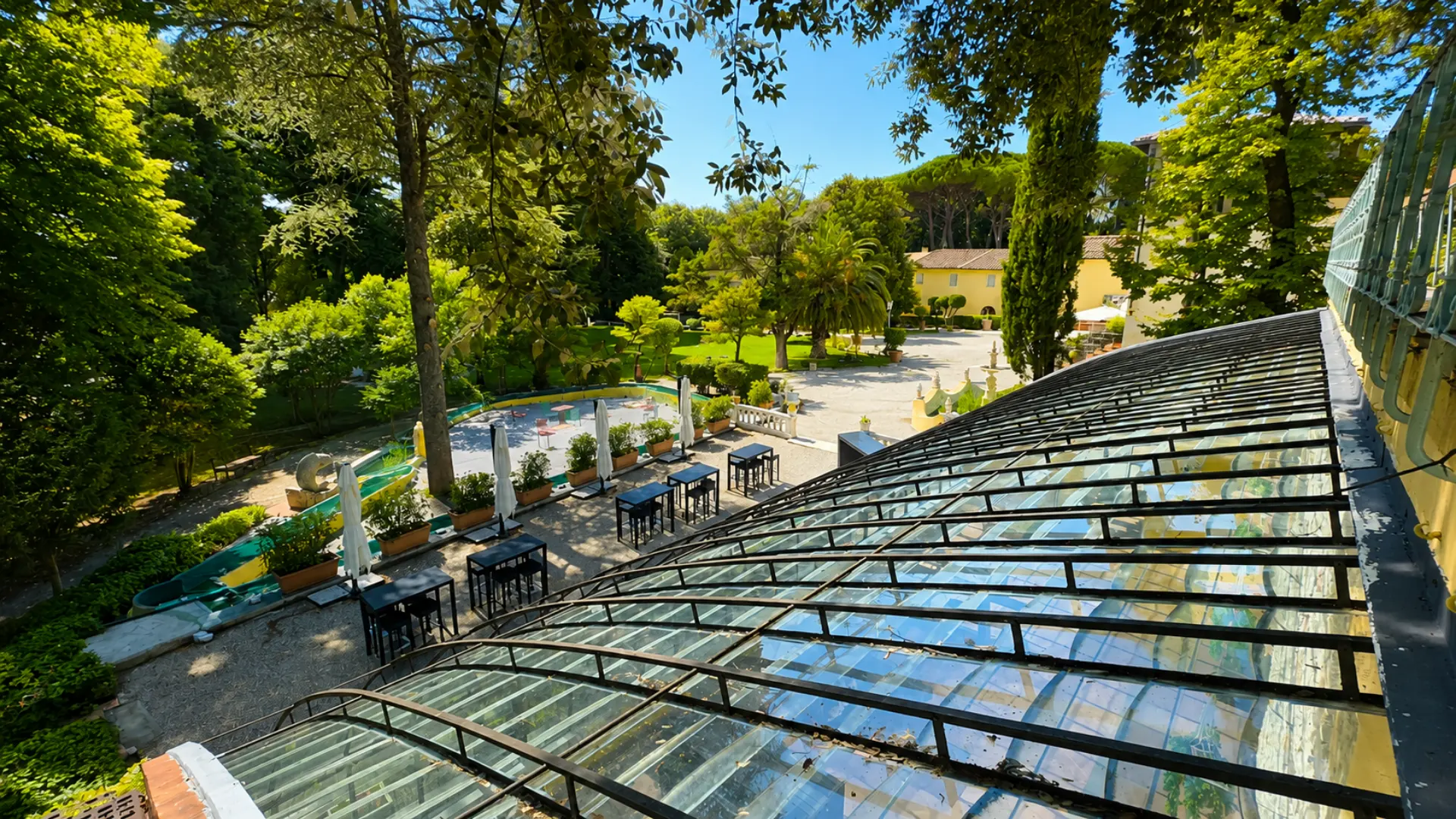 La volta a vetri vista dall'alto, e sotto la terrazza in ghiaia con i tavoli alti e gli ombrelloni chiusi, il parco e la villa sullo sfondo