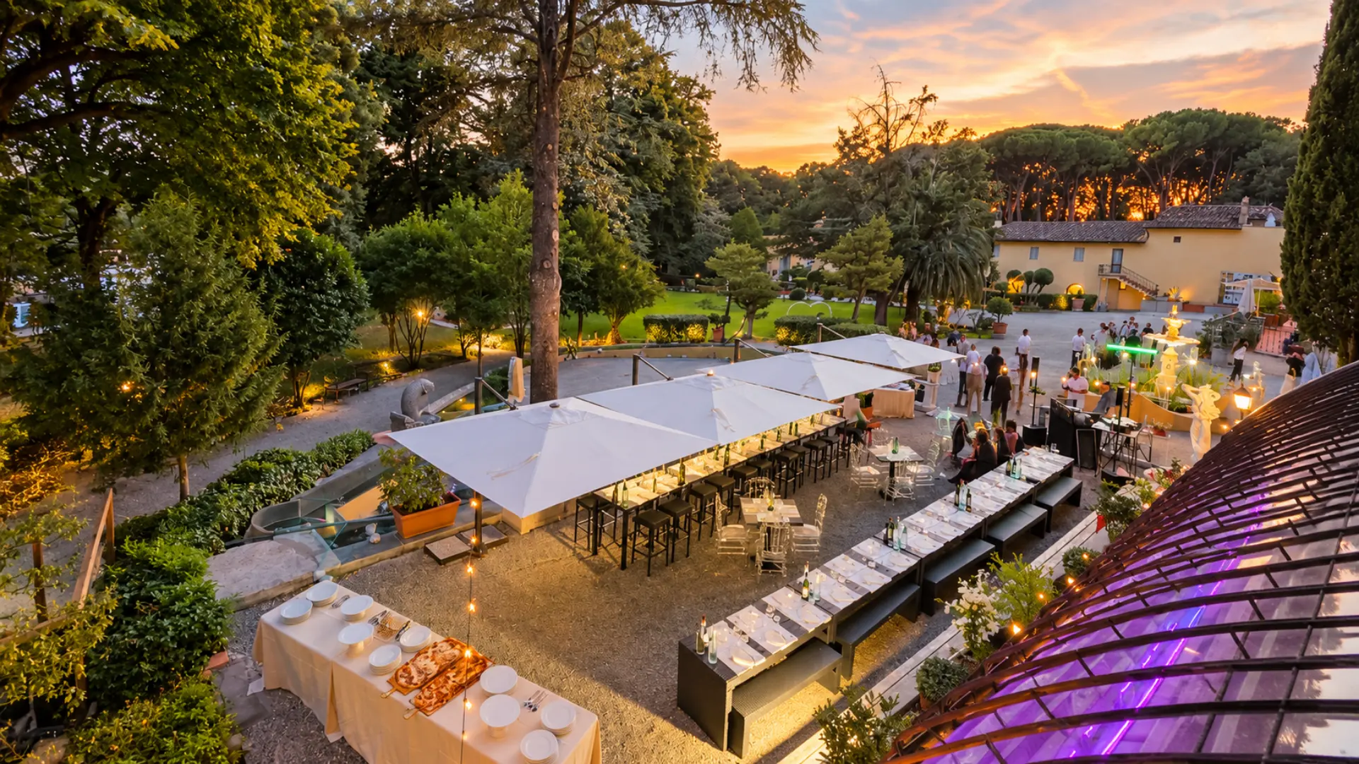 La terrazza al tramonto vista dall'alto: gli ospiti in piedi fra i tavoli, il buffet in primo piano e il cielo arancione sopra i pini